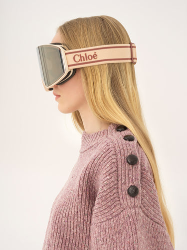 Person wearing Chloe ski goggles with a pink knit sweater on a light gray background